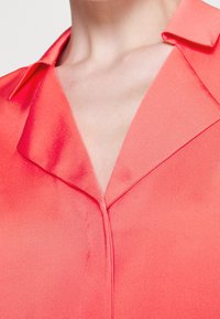 Korallfärgad satinblus med klassisk krage, djup urringning och en mjuk, glänsande yta som framhäver en smidig silhuett.