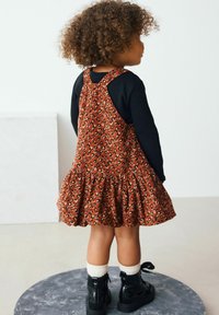 Braun gepunktetes Kleid mit Rüschensaum, getragen über einem schwarzen Langarmoberteil. Details umfassen lockiges Haar und schwarze Knöchelstiefel.