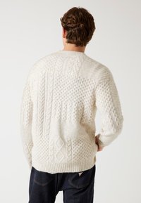 Cremfarbener gestrickter Pullover mit strukturierten Mustern, einschließlich Zopfmustern und Knötchen, mit einem gerippten Saum und langen Ärmeln.