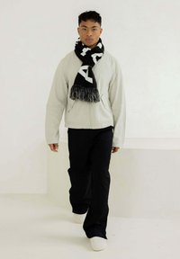 Hellgraue Zip-Jacke, schwarze Hose, weiße Schuhe und ein schwarzer Schal mit weißen Buchstaben und Fransen. Glatte Texturen und lockere Passform.