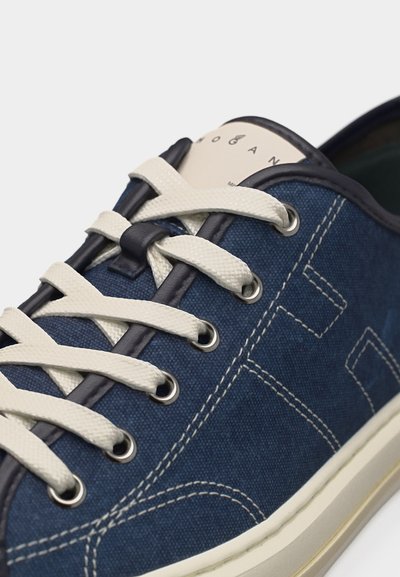 Κοντινή λήψη ενός ναυτικού μπλε πάνινο sneaker με λευκά κορδόνια, μεταλλικά οπές, σκοτεινό περιθώριο και ορατή λευκή ραφή στο πλάι.