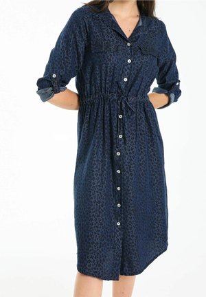 Kobieta ubrana w granatową sukienkę na guziki, długości do kolan, z podwiniętymi rękawami i paskiem ze ściągaczem w talii, z subtelnym wzorem w panterkę.