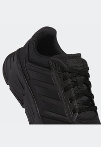 Zapatilla deportiva negra con parte superior de malla y suela de goma. Presenta tres franjas audaces en el lateral, puntera reforzada y cordones texturizados.