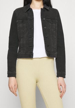 Kobieta w czarnej kurtce dżinsowej na białej koszuli i beżowych prążkowanych spodniach, stojąca na jednolitym jasnym tle.