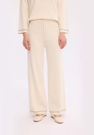 Kobieta nosząca kremowe, szerokie dzianinowe spodnie z wiązaniem w pasie i czarnym przeszyciem na lamówkach, połączone z kremowymi balerinami.