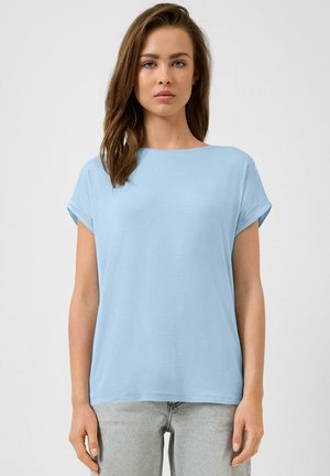 Młoda kobieta o prostych brązowych włosach, ubrana w jasnoniebieską bluzkę z krótkim rękawem i jasnoszare dżinsy, zwrócona przodem, na jednolitym tle.