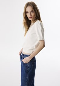 Weißes T-Shirt mit kurzen Ärmeln und runder Ausschnitt, kombiniert mit hochgeschnittenen blauen Jeans. Der Stoff sieht glatt aus und hat eine figurbetonte Silhouette.