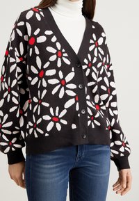Black cardigan with white and red floral pattern, featuring a V-neck, large buttons, and ribbed cuffs. Worn over a white turtleneck.