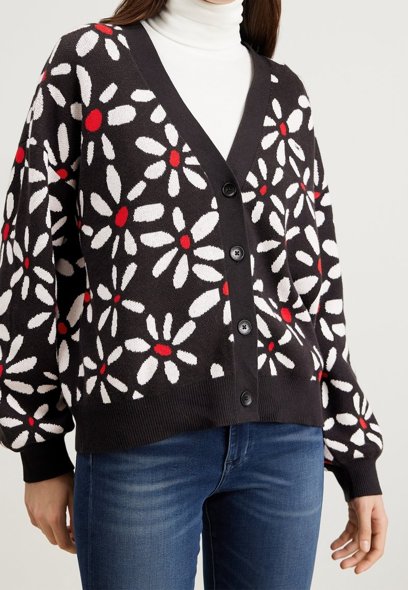 Black cardigan with white and red floral pattern, featuring a V-neck, large buttons, and ribbed cuffs. Worn over a white turtleneck.