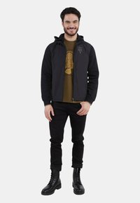 Giacca nera con cappuccio indossata sopra una t-shirt grafica verde, abbinata a pantaloni neri e stivali neri con lacci. Tessuto liscio, taglio slim, con cerniera frontale.