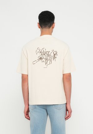 Man in een beige T-shirt met abstracte lijntekeningen van musici die instrumenten bespelen op de achterkant, gecombineerd met lichtblauwe jeans.