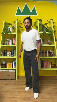 Črne kavbojke iz jeansa, bela majica in beli supergi. Prikazani pred živahno rumeno steno z zelenimi dekorativnimi poudarki in policami.