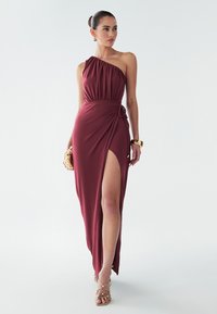 Bordové jednoloďové šaty s drapovaným živôtikom, vysokým rozparkom a detailom na viazanie na boku. Hladká látka s voľnou siluetou, v kombinácii s pásikovými sandálmi.