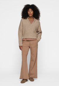 Suéter de punto marrón claro con cuello en V, diseño corto y dobladillo acanalado, combinado con pantalones beige de pierna ancha y calzado en tonos neutros.