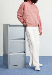 Archivador de metal gris con tres cajones, sudadera rosa, pantalones blancos con detalles rojos a los lados y zapatillas blancas sobre una alfombra azul.