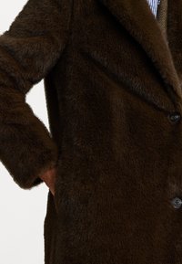 Smešani kaput od smeđeg umjetnog krzna s mekanom teksturom, s notiranim reverima, zatvaranjem na jedan gumb i bočnom džepom. Ima opušten fit.