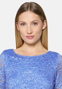 Haut en dentelle bleue avec motifs floraux, encolure ronde et manches longues. Le tissu présente une texture avec des détails complexes.