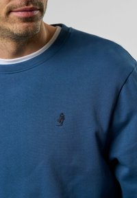 Kék pamut pulóver kerek nyakkivágással, hímzett logóval a mellkason. Simára szőtt, textúrált anyag, diszkrét varrásokkal.