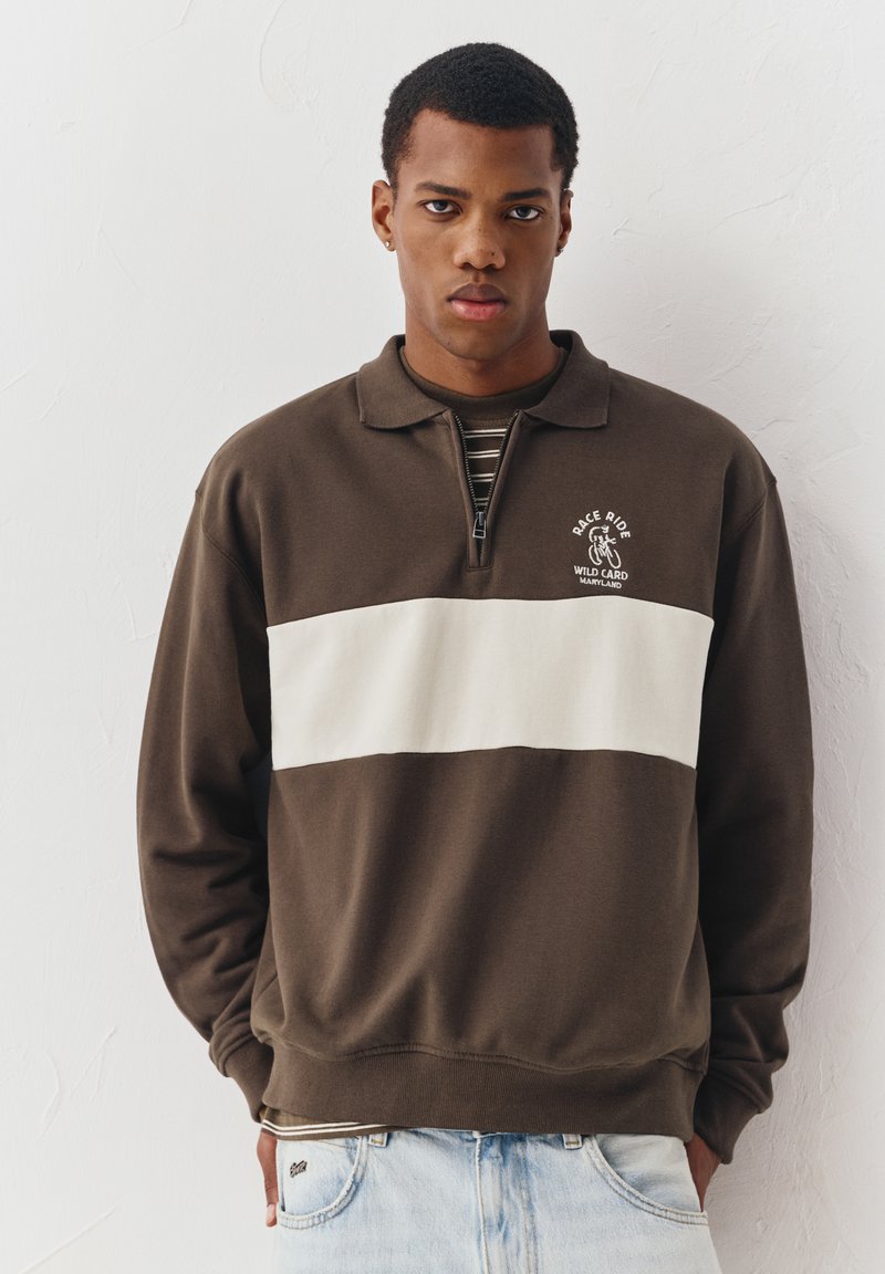 Jeune homme portant un sweat-shirt marron et blanc à fermeture éclair jusqu'au quart avec le logo "Race Ride Wild Card Maryland", debout contre un mur blanc texturé.