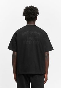Schwarzes Baumwoll-T-Shirt mit Rundhalsausschnitt, kurzen Ärmeln und dem Grafikdruck "CREATIVE DEPT. RETERNITY" auf dem Rücken.