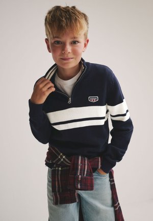 Tumši zils džemperis ar baltām svītrām, ceturtdaļas rāvējslēdzēja apkakli un logotipa plāksteri. Valkāts pāri baltai kreklam, kombinēts ar gaiši zilām džinsu biksēm un pie vidukļa sasietu rūtainu kreklu.