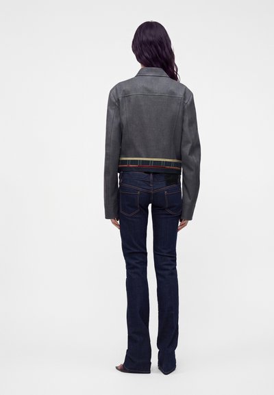 Femme aux longs cheveux foncés portant une veste courte grise et un jean évasé bleu foncé, debout de dos sur un fond blanc uni.