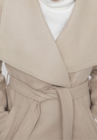 Beige ullblandad kappa med breda slag, midjebälte och sidofickor. Har en mjuk textur och slät yta. Lämplig för kallt väder.