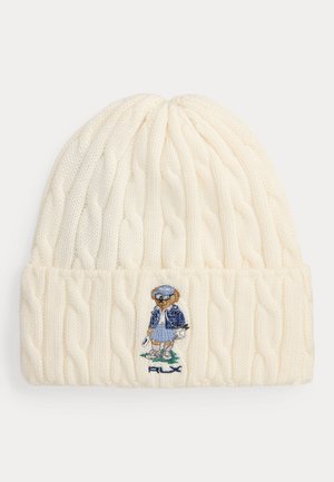 Bonnet blanc tricoté avec motif torsadé et petit ours brodé portant une tenue bleue sur le bord.
