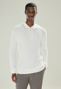 Homme portant un polo zippé à manches longues blanches texturé et un pantalon gris, se tenant avec une main dans sa poche contre un fond uni.