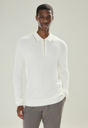 Hombre vestido con una camisa de polo blanca de manga larga texturizada y pantalones grises, de pie con una mano en el bolsillo frente a un fondo liso.