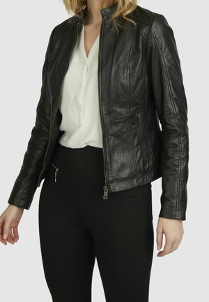 Mujer con chaqueta de cuero negra sobre una blusa blanca y pantalones negros, mostrada desde el cuello hasta las caderas sobre un fondo sencillo.