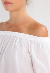 Femme portant un haut blanc à épaules dénudées, mettant en valeur l'épaule et la clavicule contre un fond clair.