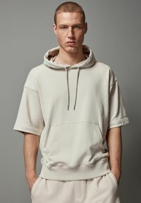 Lys beige oversized hoodie i blødt stof, med en frontlomme, snoretræk i hætten og korte ærmer. Glat tekstur med en afslappet pasform.