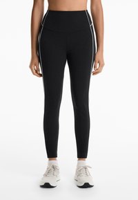 High-waisted black leggings made of stretchy fabric, featuring white side piping and a fitted design, paired with light-colored sneakers.