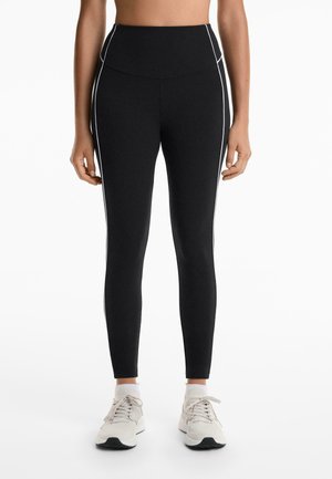 Leggings negri cu talie înaltă, realizate dintr-un material elastic, cu detalii albe pe laterale și o croială ajustată, asortați cu adidași de culoare deschisă.