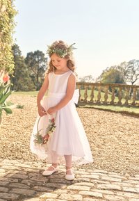 Ragazza giovane in abito bianco e corona di fiori, che tiene un cerchio floreale, in piedi su un sentiero di pietra in un giardino soleggiato.
