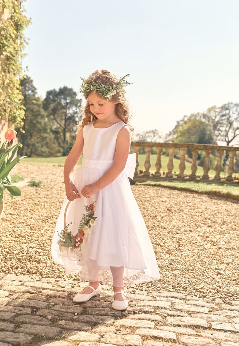 Ragazza giovane in abito bianco e corona di fiori, che tiene un cerchio floreale, in piedi su un sentiero di pietra in un giardino soleggiato.