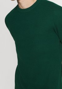 Homme portant un pull en maille texturée vert foncé avec un col rond, visible de l'épaule à la taille sur un fond uni.