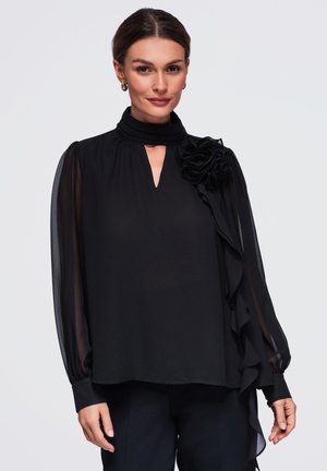 Femme portant un chemisier noir à col montant avec des manches bouffantes transparentes et une grande fleur en tissu sur l'épaule gauche.