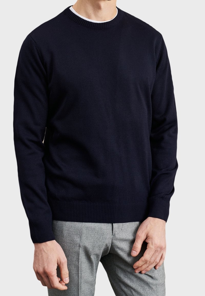 Navy crew-neck sweater in a soft knit fabric with ribbed cuffs and hem. Paired with grey checked trousers for contrast.