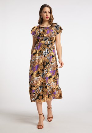 Frau trägt ein bunt gemustertes Midikleid mit lila, schwarzen, goldenen und weißen Designs, kombiniert mit braunen Sandalen mit Knöchelriemen.