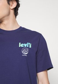 Gros plan d'une personne portant un t-shirt Levi's bleu marine avec un logo vert et un motif de vague blanche au-dessus du texte « California ».