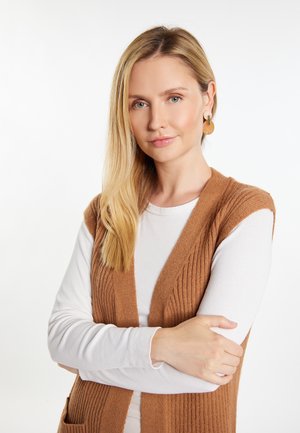 Canotta a coste marrone sopra una camicia bianca a maniche lunghe, con scollo a V e tasche laterali, con capelli lisci e dritti e orecchini circolari.