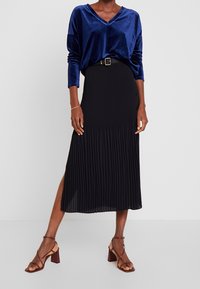 Top in velluto blu navy a maniche lunghe con scollo a V; gonna midi plissettata nera con spacco laterale e bottoni laterali; sandali con tacco strappy borgogna.