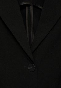 Sort blazer med en notch lapel, enkelt sort knaplukning og glat tekstur. Indersiden viser mørkt stoftrim langs kanten.