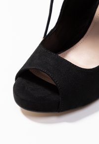 Czarne zamszowe buty na wysokim obcasie z odkrytą czubkiem i paskiem wokół kostki, z widoczną szczegółową strukturą i beżowym wnętrzem na białym tle.