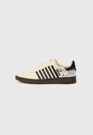 Cream leather sneaker with black stripes, brown sole, and metallic silver heel. Features a logo on the side and classic lace-up design.