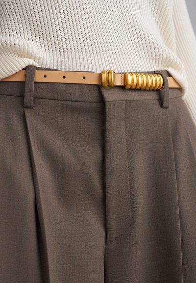 Pantaloni marroni a pieghe con finitura testurizzata, abbinati a una cintura beige con accenti circolari dorati e multiple aperture di regolazione.