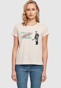 Lys pink bomulds t-shirt med sort tryk af en figur, der holder en malerpensel, og grøn tekst, der siger "FØLG DINE DRØMME", overstreget med rød.