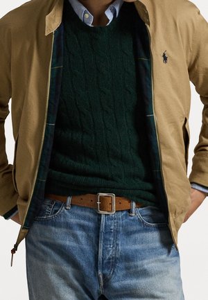 Hombre con chaqueta marrón claro sobre suéter de punto grueso verde oscuro, camisa azul de botones, cinturón marrón y vaqueros azules, con las manos en los bolsillos.